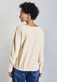 Maglia a maniche lunghe, di un beige chiaro, con una vestibilità morbida, scollatura rotonda e orlo arricciato. Abbinata a jeans in denim blu. Il tessuto ha una consistenza morbida.