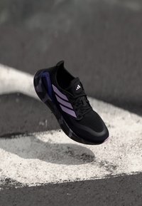 Chaussure de sport noire avec une tige en maille, des accents violets et trois bandes blanches. Elle présente une semelle texturée et un design léger.