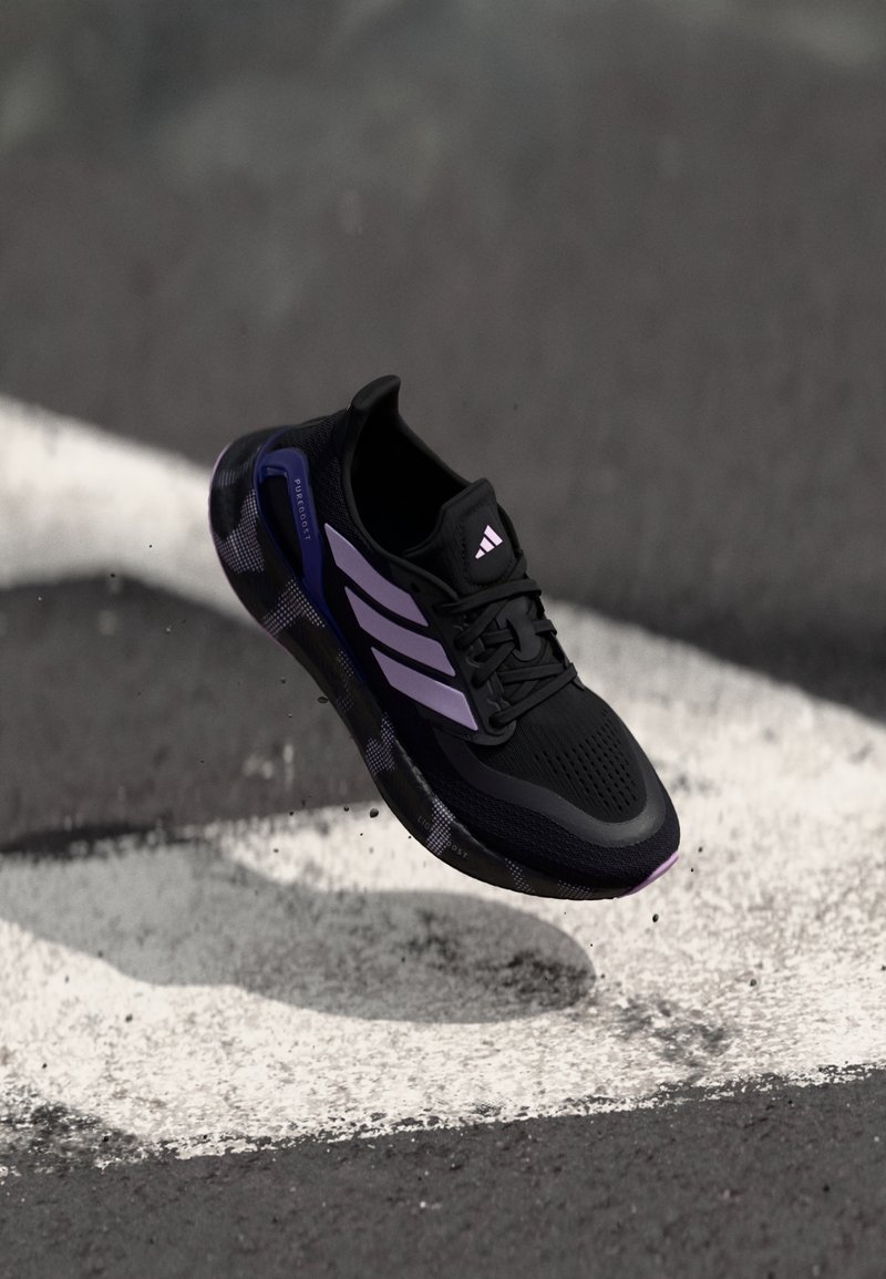 Chaussure de sport noire avec une tige en maille, des accents violets et trois bandes blanches. Elle présente une semelle texturée et un design léger.