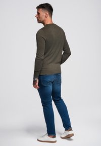 Maglietta a maniche lunghe verde oliva con texture a coste, abbinata a jeans blu a gamba dritta e sneaker bianche, vista da dietro.