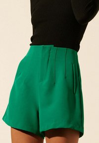 Shorts en satin vert avec une texture lisse, dotés d'une taille haute, de poches latérales et de plis subtils. Associés à un haut noir à côtes.