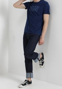 Tmavě modré tričko s kulatým výstřihem a grafickým motivem, kombinované s tmavými džínami s ohrnutým lemem a černými teniskami s bílými detaily.