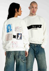 Weißer Sweatshirt mit grafischen Patches, die Porträts und Text zeigen. Mit gerippten Bündchen und einer weichen Textur, kombiniert mit lockeren blauen Jeansshorts.