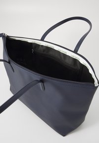 Sac fourre-tout bleu marine en matériau texturé, avec une fermeture éclair et deux longues poignées. L'intérieur est orné d'une bordure en tissu de marque et d'une poche zippée.