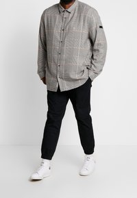 Camisa de cuadros gris y negra con cuello, combinada con pantalones jogger oscuros y zapatillas blancas. Botones destacados y etiqueta en la manga.