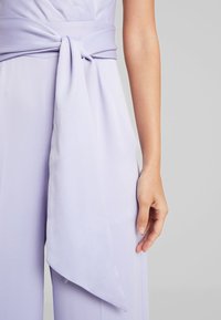 Lila jumpsuit med knytning i midjan. Den släta texturen, de vida benen och den draperade silhuetten förstärker den övergripande designen.