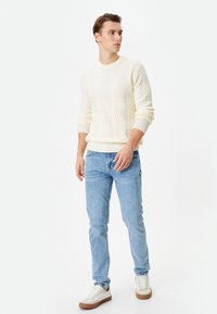 Suéter de punto crema con un patrón de espiga texturizado, combinado con jeans azul claro y zapatillas blancas con suelas marrones.