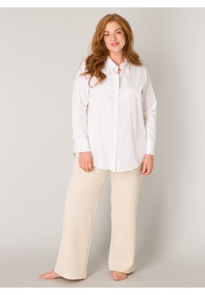 Vrouw met lang rood haar, gekleed in een wit overhemd met knopen en beige brede broekspijpen, staand op blote voeten tegen een lichtgrijze achtergrond.