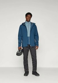 Άνδρας με στρωματοποιημένα μπλε μπουφάν και σκούρους παντελόνες, κρατώντας ένα μαύρο κράνος ποδηλάτου, ενώ στέκεται μπροστά σε απλή ανοιχτόχρωμη πρόβα.