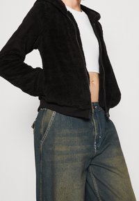 Felpa nera in pile con zip e superficie testurizzata. Presenta un cappuccio e polsini a coste, abbinata a jeans a gamba larga blu scoloriti.