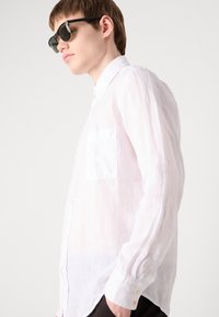 Camisa de lino blanca con mangas largas, un bolsillo en el pecho y botones en el frente. El modelo lleva gafas de sol negras y pantalones oscuros.