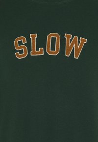 Grünes Baumwollhemd mit dem gestickten Wort "SLOW" in Braun und strukturiertem Finish, vor einem glatten Stoffhintergrund.