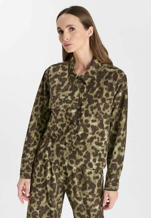 Frau mit langen braunen Haaren, die eine grün-braune, knöpfbare Jacke mit Leopardenmuster und passende Hosen vor einem schlichten Hintergrund trägt.
