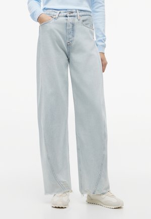 Isik, kes kannab helerohelisi laiade säärtega teksapükse, millel on lahtised ääred, valgeid tenniseid ja helerohelist pikema varrukaga särki, seisab ühe käega taskus.