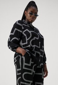 Camisa negra de botones y pantalones a juego con patrones geométricos blancos. La tela tiene una textura suave, un corte holgado y mangas enrolladas.