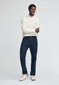 Beige Pullover mit Rundhalsausschnitt und schwarzem Logo, kombiniert mit dunkelblauen Jeans und hellen Sneakers. Schlichtes Design, figurbetonte Passform und lässiger Stil.