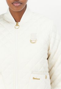Gepolsterte cremefarbene Jacke mit einem Reißverschluss, goldenen Beschlägen, elastischen Bündchen und einer Vordertasche mit Logodetail. Strukturierter Stoffdesign.
