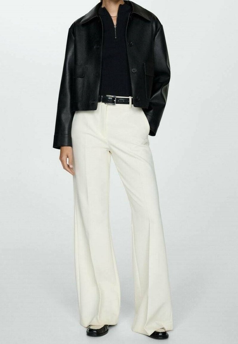 Personne portant une veste noire sur un haut noir, un pantalon blanc à jambes larges, une ceinture noire et des chaussures noires, debout devant un fond uni.