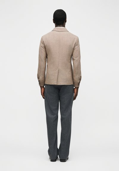 Άνδρας με ρούχα από πίσω, φοράει μπεζ σακάκι με πίσω σκίσιμο και σκουρόχρωμο γκρι παντελόνι, στέκεται μπροστά σε απλό ανοιχτό φόντο.