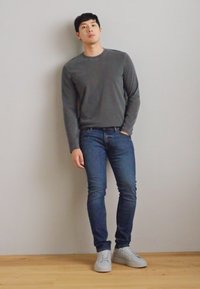 Camisa de manga longa cinza, jeans azul e ténis brancos. A camisa tem um pescoço redondo; os jeans são de corte justo com um design clássico.