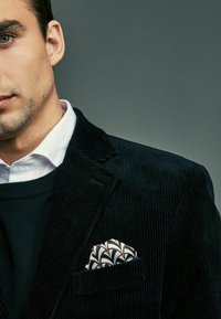 Mornarsko plavi kord blazer složen preko crnog pulovera, sa bijelim ovratnikom košulje i šarenim maramicom s akcentima u crnoj i smeđoj boji.