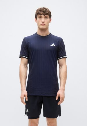 Chemise de sport bleu marine avec des rayures verticales, manches courtes et accents blancs. Associée à un short noir avec un logo.
