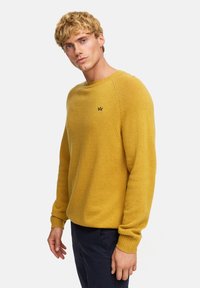 Musztardowy sweter dzianinowy z długimi rękawami, żebrowanymi mankietami i okrągłym dekoltem, z małym czarnym logo na piersi.