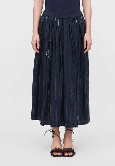 Femme portant une jupe midi plissée bleu marine avec des rayures verticales en sequins et des sandales noires à bouts ouverts avec des liens à la cheville.