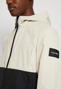Witte en zwarte lichte jas met een ritssluiting aan de voorkant, capuchon, zijzakken en een zwart Calvin Klein-logopatch op de linker mouw.