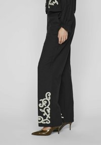 Pantalones rectos negros con intrincado bordado floral blanco en el dobladillo, combinados con zapatos de tacón marrones de punta. Textura de tejido suave.