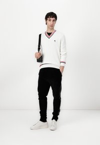 Weißer Strickpullover mit roten und schwarzen Akzenten, schwarze Cord-Jogginghosen und weiße Sneaker. Das Modell hält eine kleine schwarze Tasche.