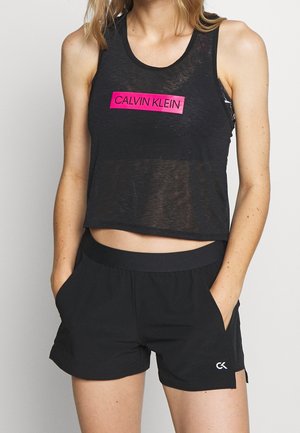 Žena v černém krátkém topu s nápisem "Calvin Klein" růžovou barvou a černých šortkách s logem "CK", ruce v kapsách.