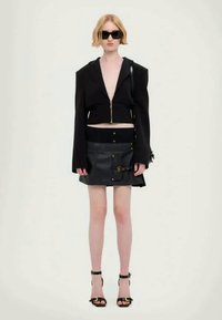 Fekete, rövidített dzseki cipzárral, fekete bőr mini szoknyával, arany kiegészítőkkel párosítva. A modellen fekete napszemüveg és magassarkú cipő van.