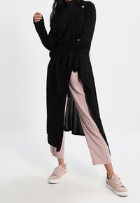 Long gilet noir avec devant drapé et ceinture à nouer, porté sur un haut noir et un pantalon large côtelé rose clair. Des baskets rose clair complètent le look.