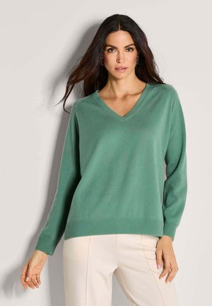 Frau mit langen, dunklen Haaren, die einen grünen V-Ausschnitt-Pullover und hellbeige Hosen trägt, steht vor einem schlichten hellen Hintergrund.