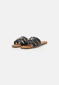 Sandales noires à glissière avec trois larges lanières, présentant un accent de chaîne dorée et une semelle plate marron. Texture en cuir lisse.