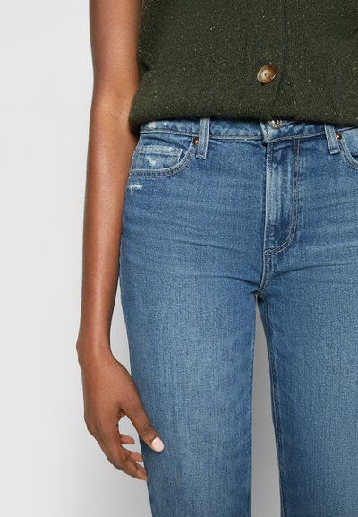 Calças de ganga azuis com cintura alta, apresentando desgaste subtil, design de cinco bolsos e uma textura suave. Usadas com um top de malha verde escuro.