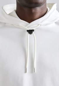 Weißer Hoodie aus strukturiertem Stoff, ausgestattet mit einem schwarzen, dreieckigen Zugstopper für die Kordeln und einem schlichten, glatten Design.