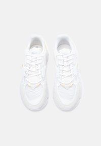 Valged sportlikud kingad, millel on võrgu ülemine osa, nahast detailid, tekstureeritud kummitald ja luku sulgemine koos kollaste aktsentidega.