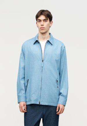 Jonge man met kort donker haar, gekleed in een lichtblauw jasje met ritssluiting over een wit shirt en donkerblauwe broek, tegen een effen lichte achtergrond.