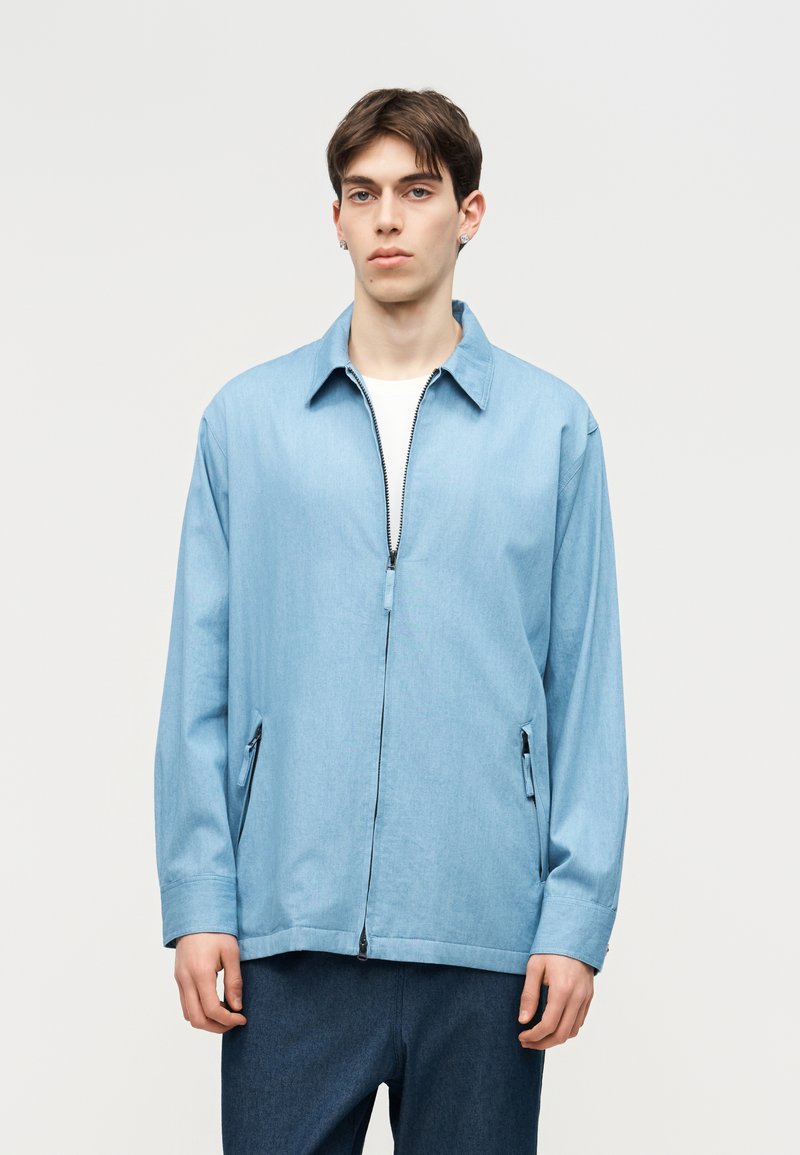 Νέος άνδρας με κοντά σκούρα μαλλιά, που φοράει ανοιχτό μπλε μπουφάν με φερμουάρ πάνω από λευκό πουκάμισο και σκούρο μπλε παντελόνι, σε απλό φωτεινό φόντο.