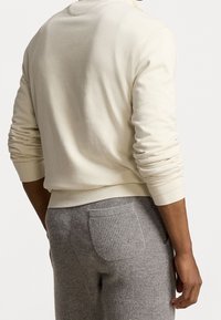Sudadera de algodón color crema con puños de canalé, combinada con pantalones grises texturizados que tienen un bolsillo trasero. Vista cercana desde la parte posterior.