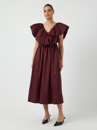 Robe bordeaux, longueur genou, avec un décolleté en V profond et de grandes manches volantes, présentant une taille cintrée et un tissu léger. Escarpins noirs à bouts pointus.