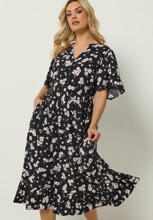 Kvinde med blondt hår iført en sort midi kjole med pink blomsterprint, V-hals, korte ærmer og flæsekant, poserer med hænderne i lommerne.