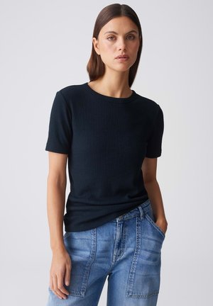 Frau mit glatt braunen Haaren, die ein schwarzes, geripptes Kurzarmshirt und blaue, hoch taillierte Jeans trägt, steht vor einem hellen Hintergrund.