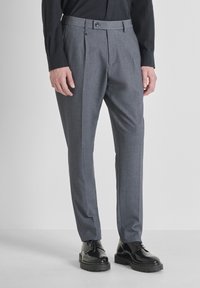 Pantaloni grigi su misura con una texture liscia, caratterizzati da pieghe frontali e chiusura con bottone, abbinati a scarpe lucide nere.