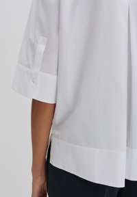 Blusa bianca con bottoni, caratterizzata da maniche corte con dettagli a bottone, un orlo dritto e un tessuto liscio e testurizzato.