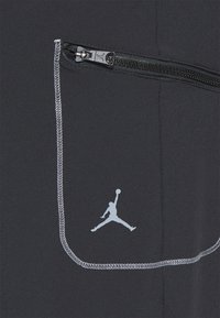 Svarta träningsbyxor med en ficka med dragkedja, med vit sömnad och en ljusgr å Jordan-logotyp på fickan. Slät yta.