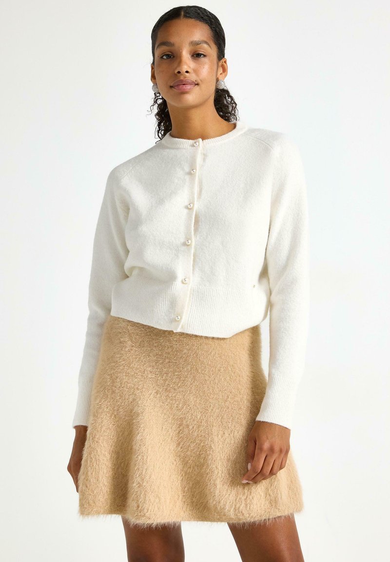 Cardigan bianco lavorato a maglia con bottoni perla abbinato a una gonna fluffy beige. Il cardigan ha orlo a coste e maniche lunghe.