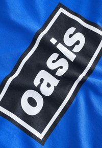 Blauwe stof met een zwart rechthoekig vlak waarop de tekst "oasis" in vetgedrukte witte letters staat. De textuur lijkt glad en licht reflecterend.